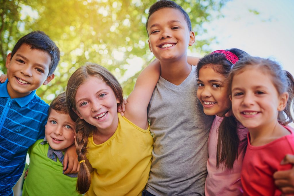 Six young children with their arms around each other, looking at the camera and smiling. They are practicing good social skills.
