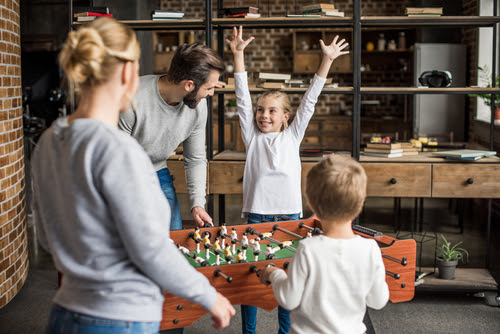 Family around a foosball table, the young daughter celebrating by throwing her hands in the air.