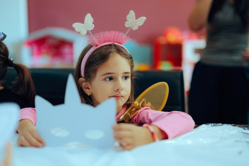 A young girls in a costume sitting at a table and holding a mask.
